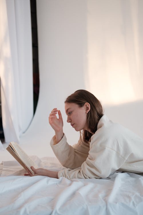 Free Side view of young female reading book and pondering while relaxing on soft mattress in light room Stock Photo