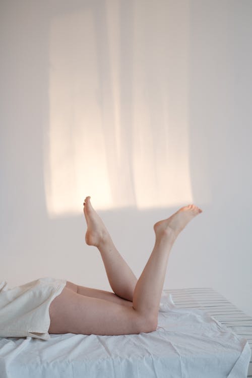 Side view of anonymous sensual woman in shirt swinging barefoot legs while relaxing on soft mattress against white wall