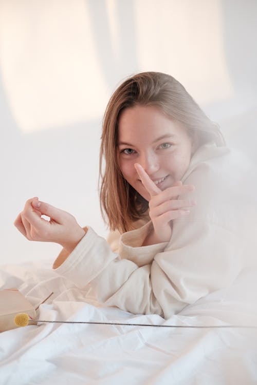 Free Positive young woman looking at camera with smile and touching lips with finger while relaxing on soft mattress in morning at home Stock Photo