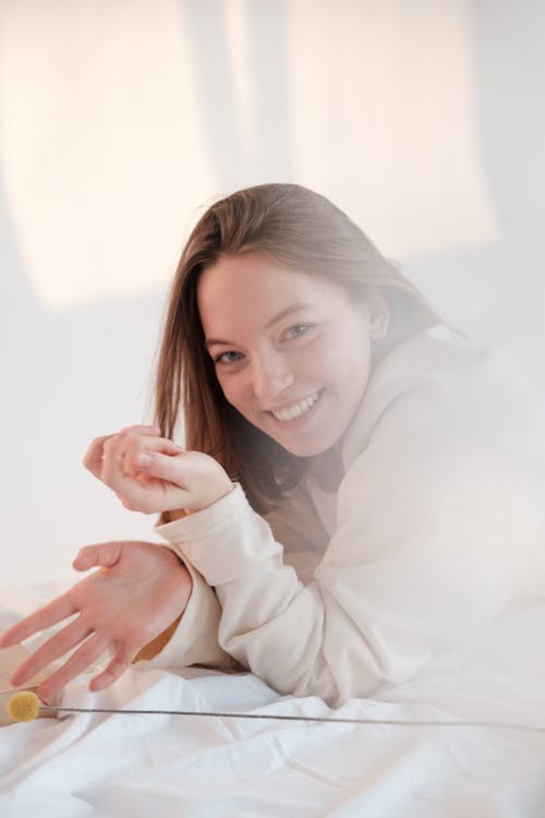 Free Cheerful young woman smiling for camera and touching flower while relaxing on bed in morning at home Stock Photo