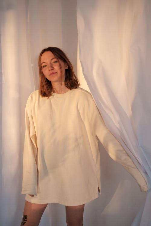 Free Young female in sweatshirt looking at camera while standing near white curtains at home Stock Photo