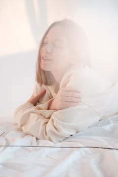 Young female with closed eyes touching shoulders and enjoying softness of sweatshirt while resting on bed in morning