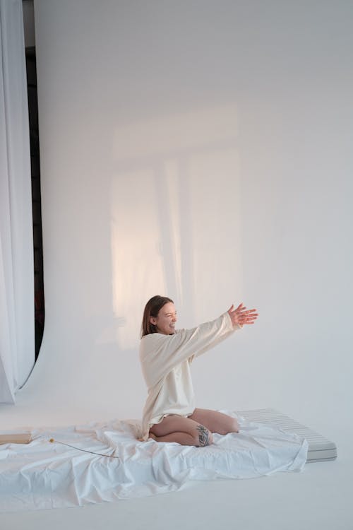 Free Delighted young female in sleepwear smiling and stretching out arms to touch sunlight while sitting on soft mattress against white background in morning Stock Photo