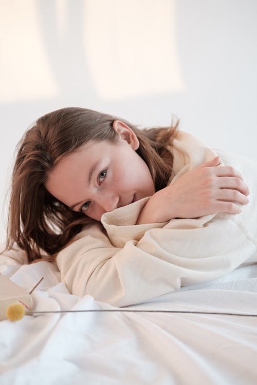 Young woman in soft sweatshirt touching shoulders and looking at camera while relaxing on bed near flower