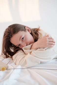 Young woman in soft sweatshirt touching shoulders and looking at camera while relaxing on bed near flower