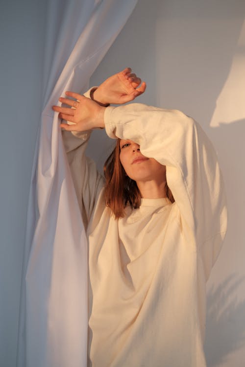 Young female in sweatshirt covering face with raised arms and looking at camera while standing near white curtain in sunlight