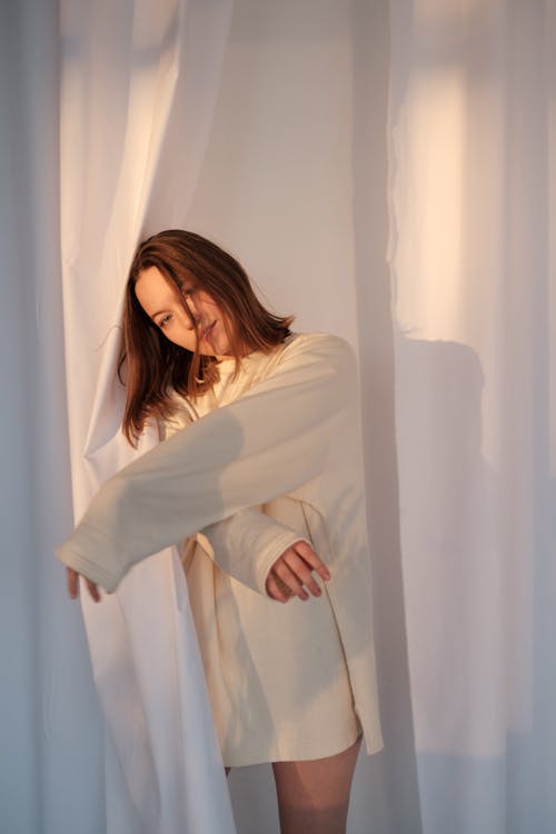 Free Young woman in sweatshirt leaning on white curtain and looking at camera in light room Stock Photo