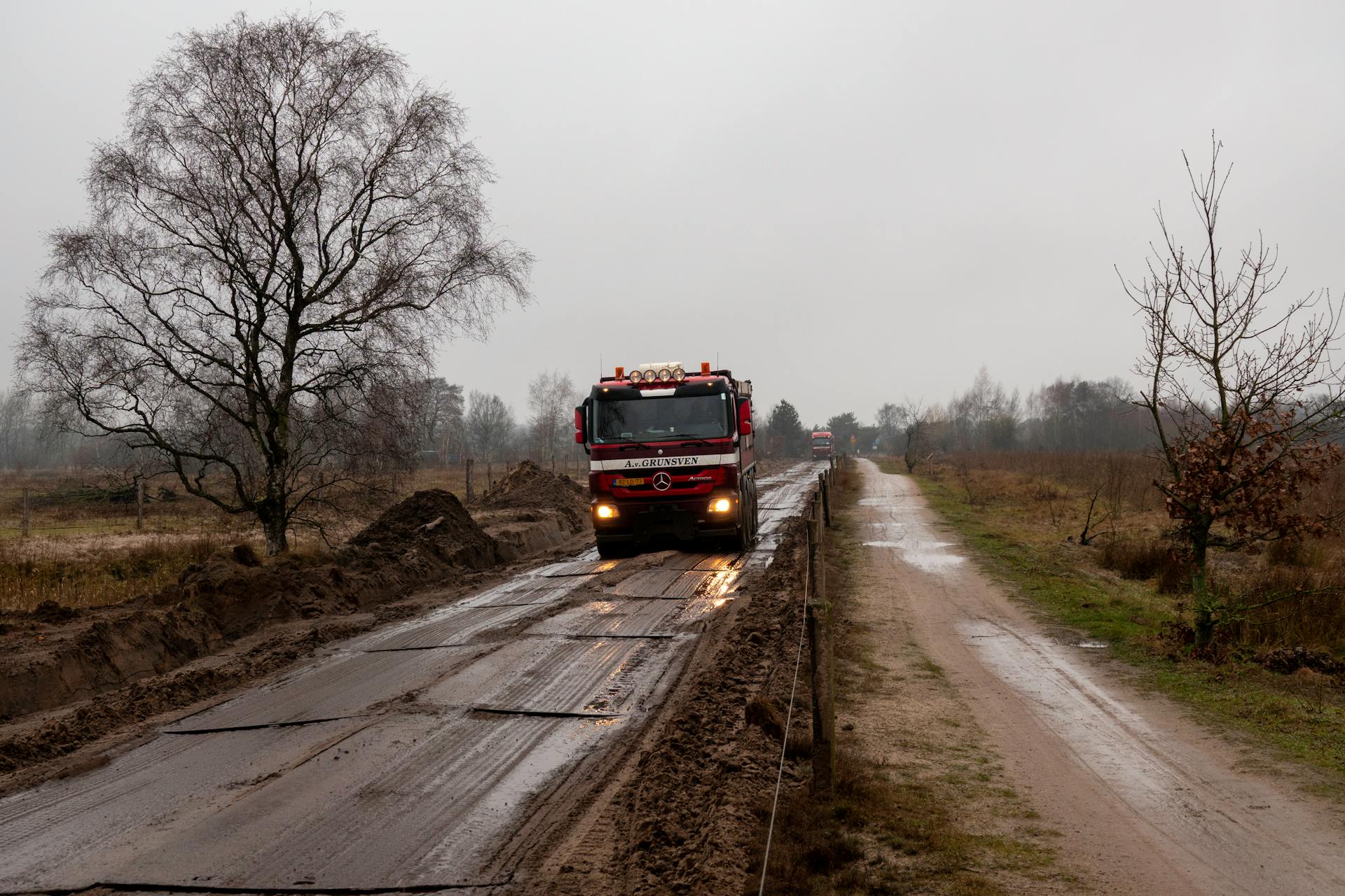 Red truck navigating a muddy road in the Netherlands countryside on a gloomy day.