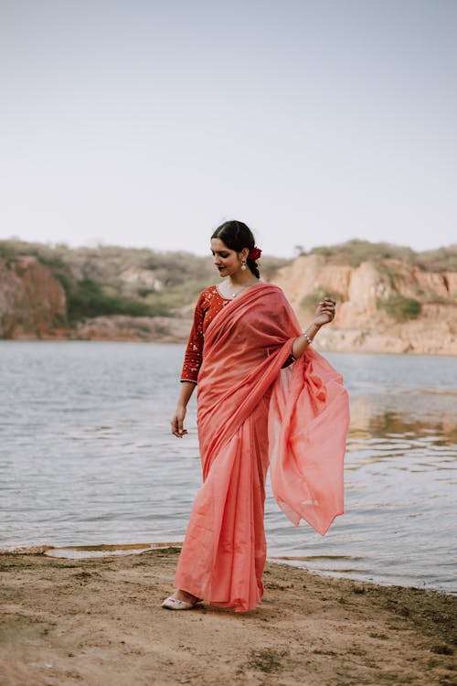 Full body of young Indian female wearing light red sari standing on sandy coastline at calm ripple of river