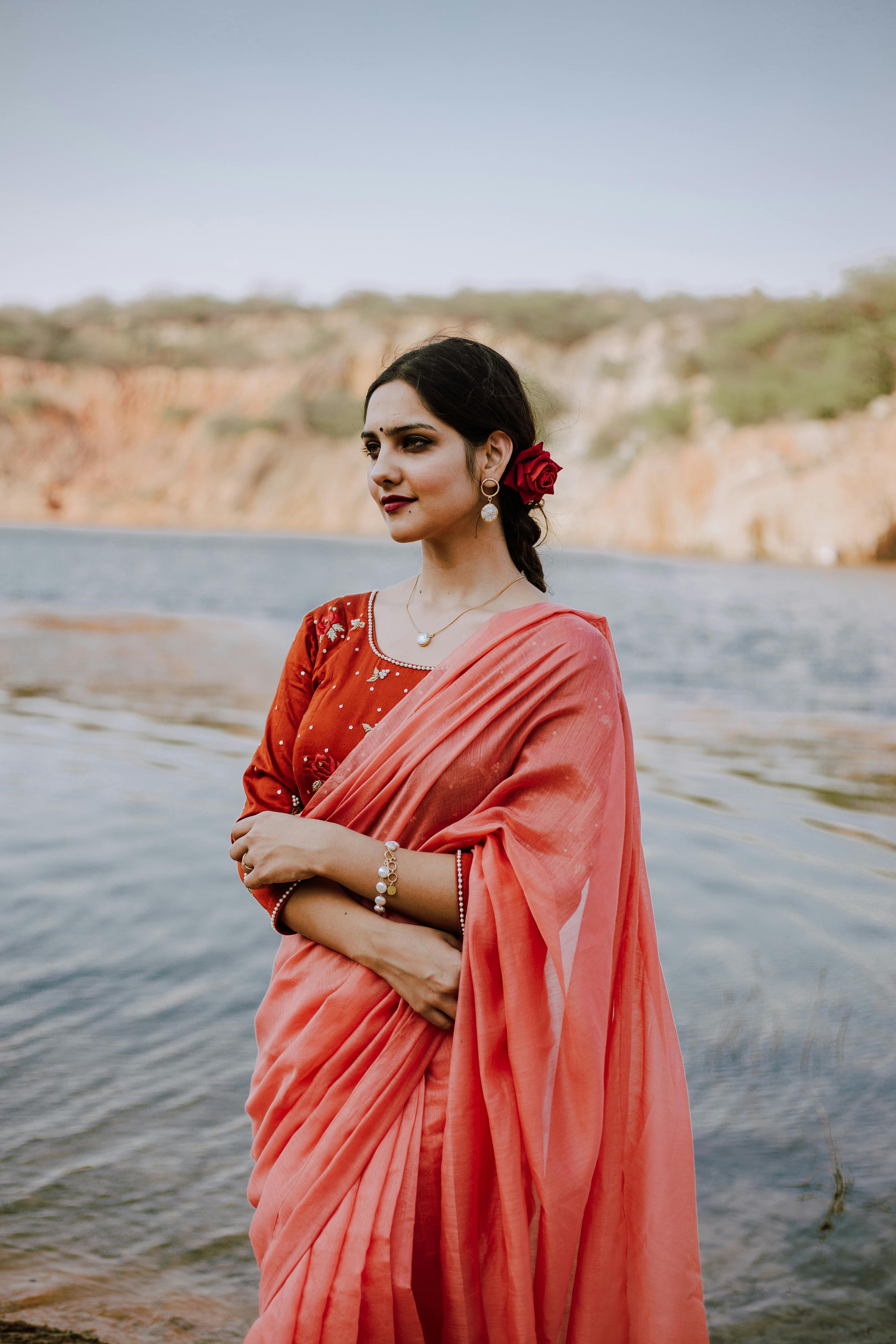 Positive young ethnic female wearing traditional Indian sari standing with crossed arms on waistline on shore of river