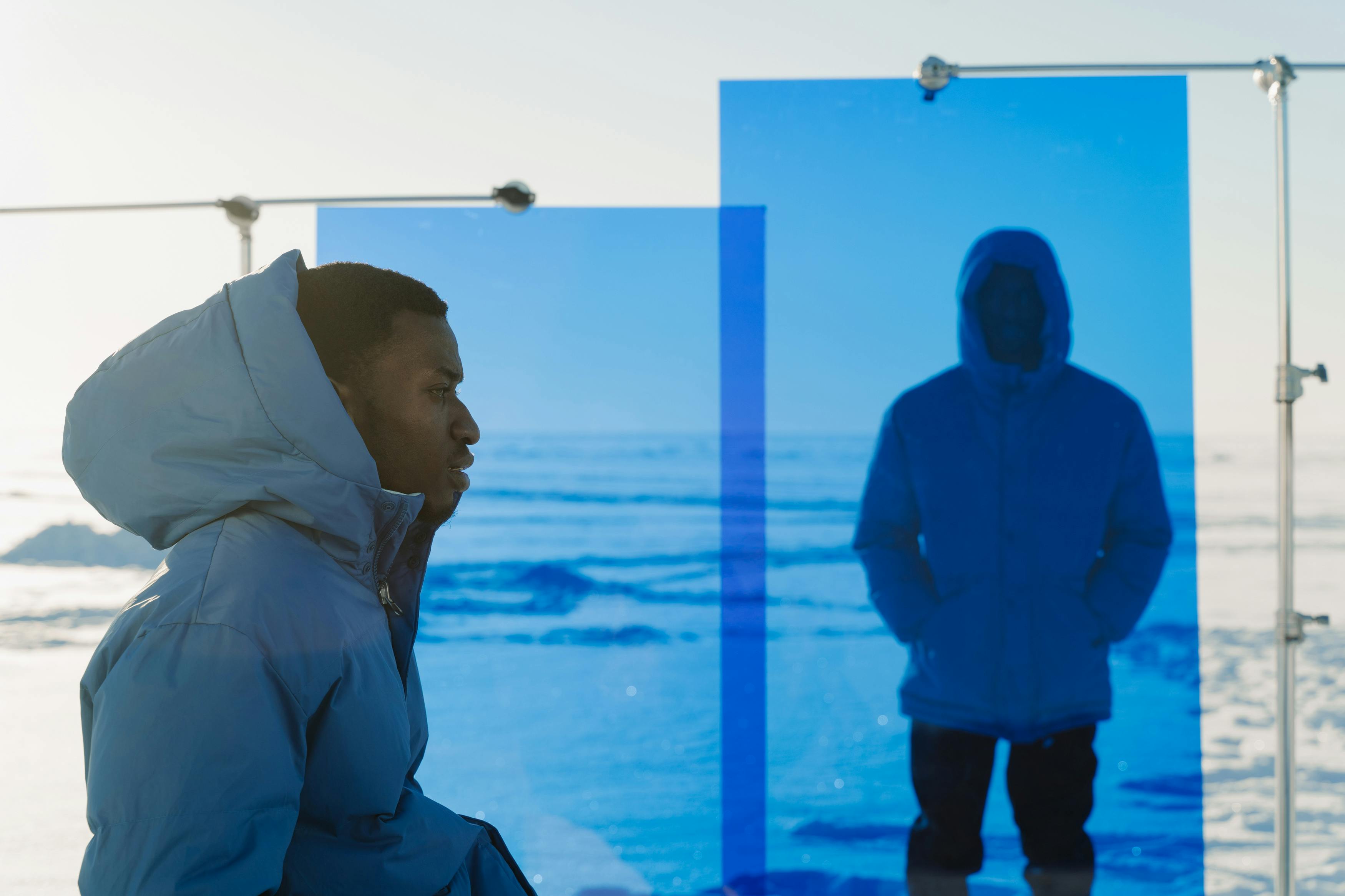 Man in blue puffer jacket reflected in glass with snowy background.