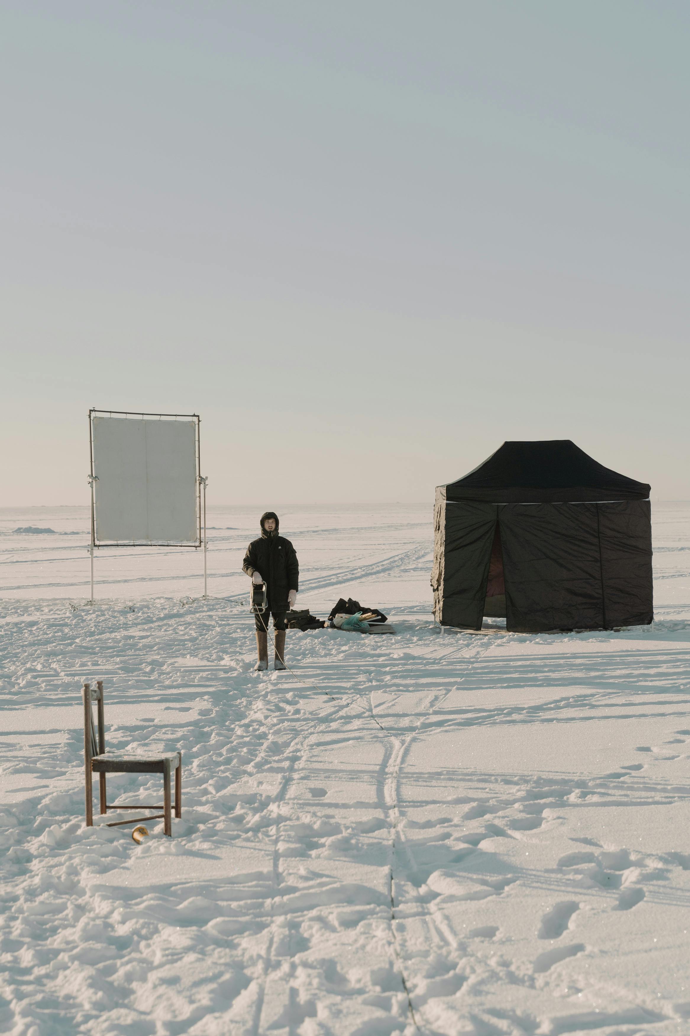 A winter camping scene featuring a tent and chair on snow-covered ground with a person standing nearby.