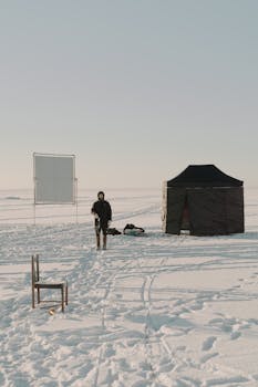 A winter camping scene featuring a tent and chair on snow-covered ground with a person standing nearby.