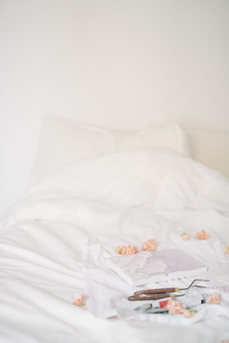 A Notebook Beside Painting Materials Surrounded By Petals On A White Blanket