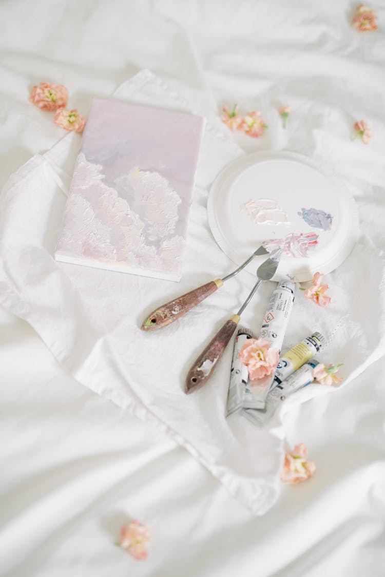 A Notebook Beside Painting Materials Surrounded By Petals On A White Blanket