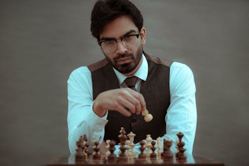 Concentrated masculine bearded ethic male wearing formal clothes sitting at board and making chess move on blurred gray background