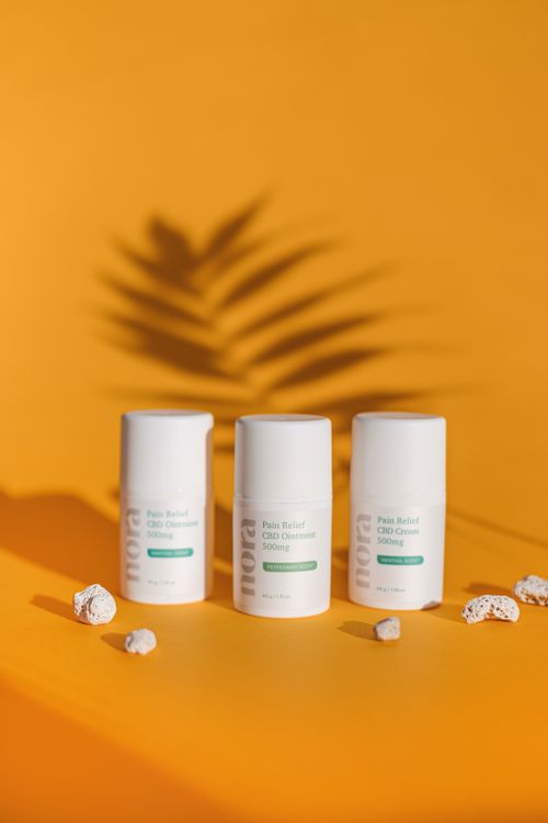 Free Three bottles of pain relief CBD cream on a vibrant orange surface with a shadowed leaf pattern. Stock Photo
