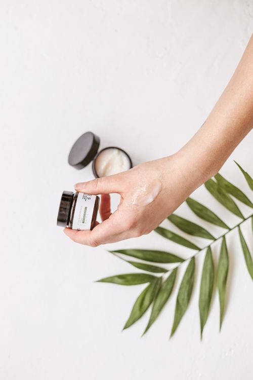Free Hand holding a skincare cream jar next to a green leaf on a white background. Stock Photo