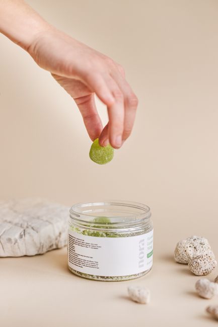 A hand holding a green gummy above an open jar on a minimalist background.