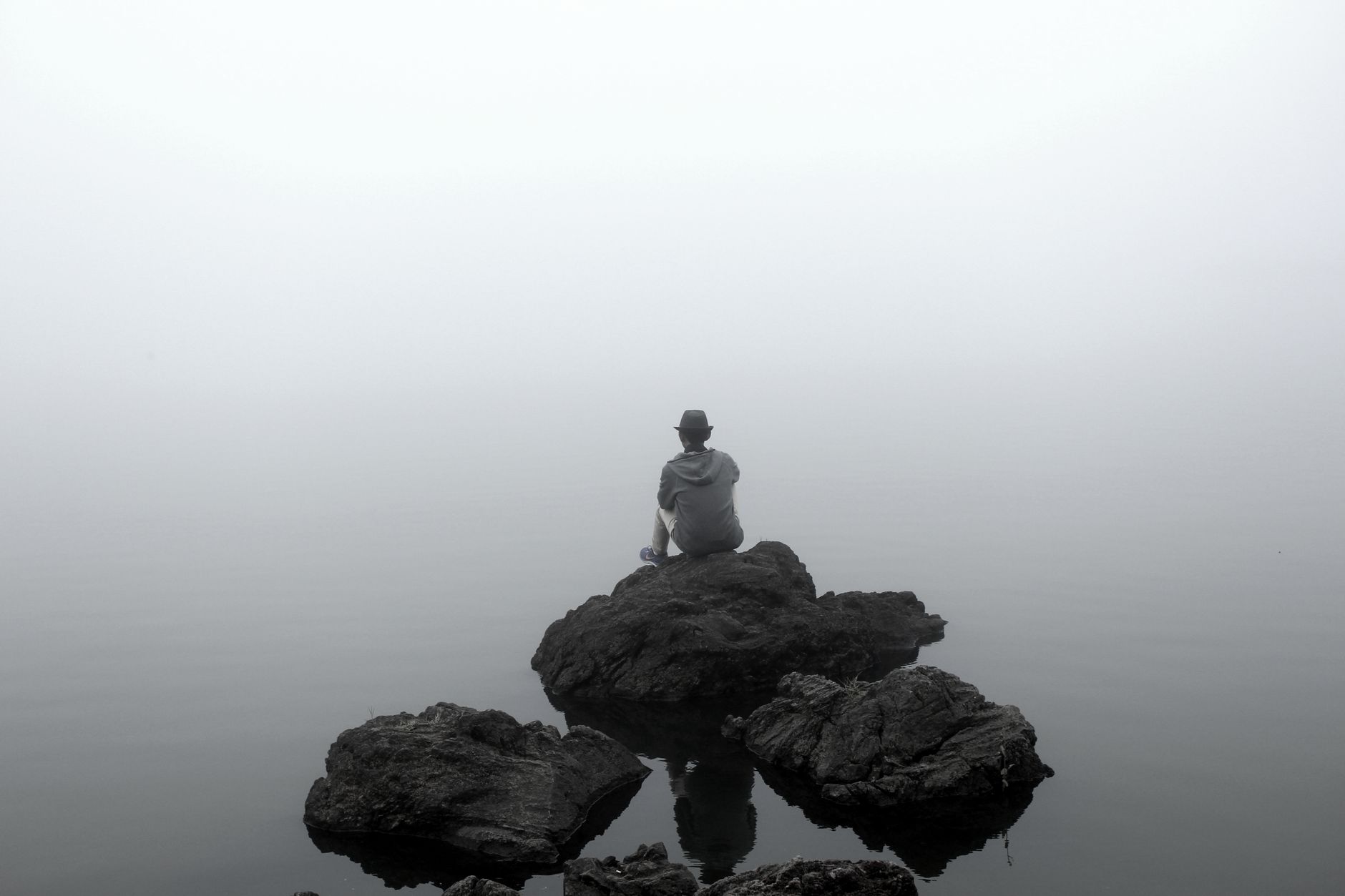 A person sits alone on rocks, contemplating in a foggy, serene setting by the water.