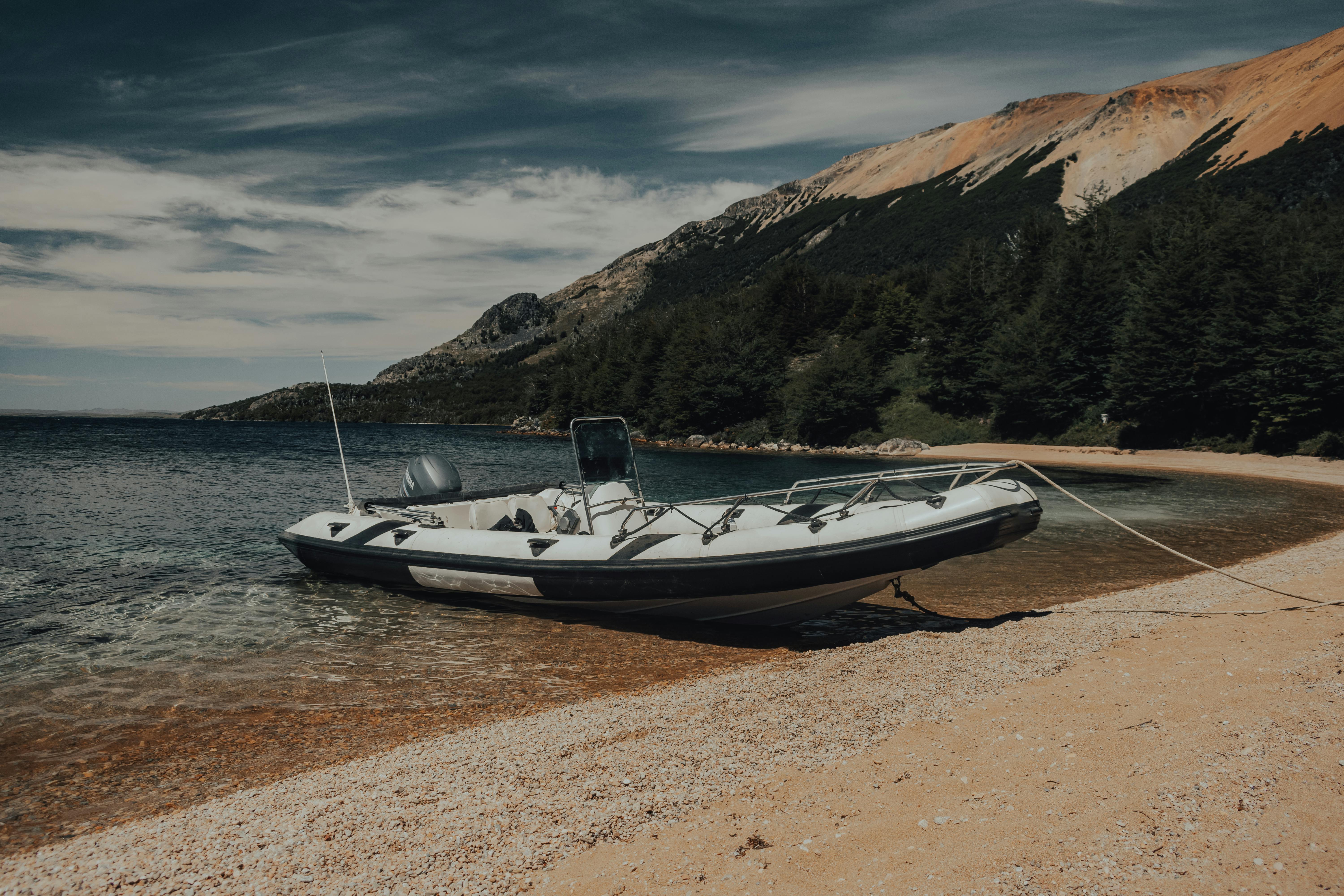 Free Inflatable boat anchored on a tranquil sandy beach beside clear waters and mountains. Stock Photo