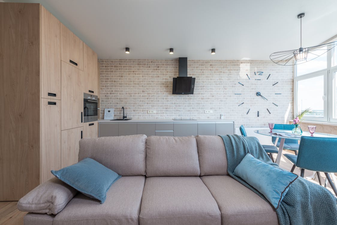 Minimalistic interior design of modern light studio consisting of big sofa and kitchen with dining table and chairs against brick wall in daylight