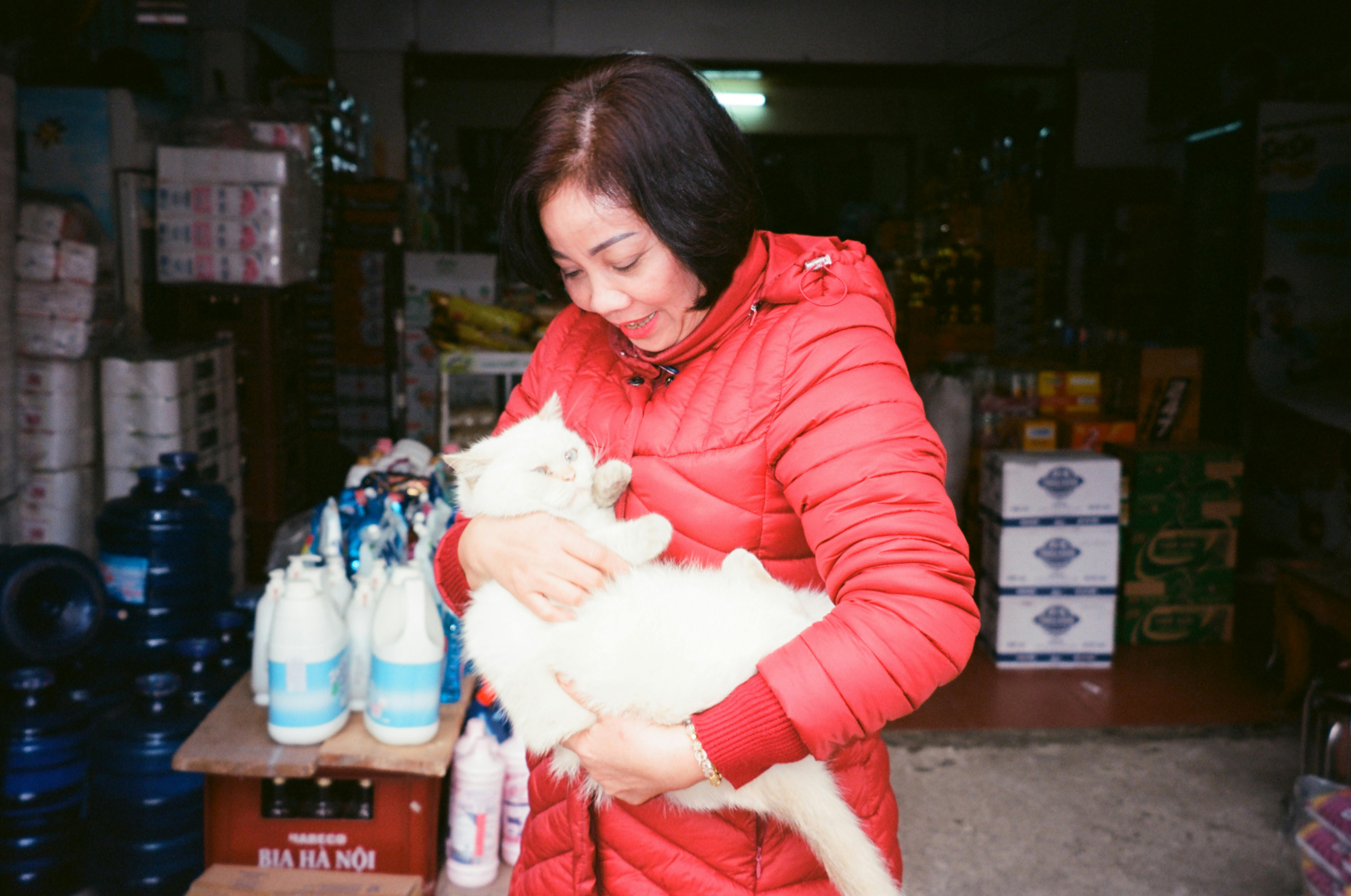 Smiling woman in a red jacket holds a fluffy white cat inside a store.