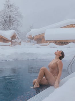 A woman relaxes by an outdoor pool surrounded by snow-covered cabins during winter snowfall.