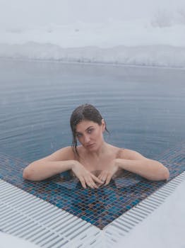 A woman relaxes in a steaming outdoor pool during a snowy winter day, creating a serene contrast.