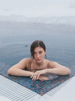 Caucasian woman swimming in a heated outdoor pool while snow falls around, creating a serene winter scene.