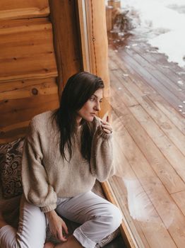 A woman in a sweater gazes thoughtfully out a cabin window.