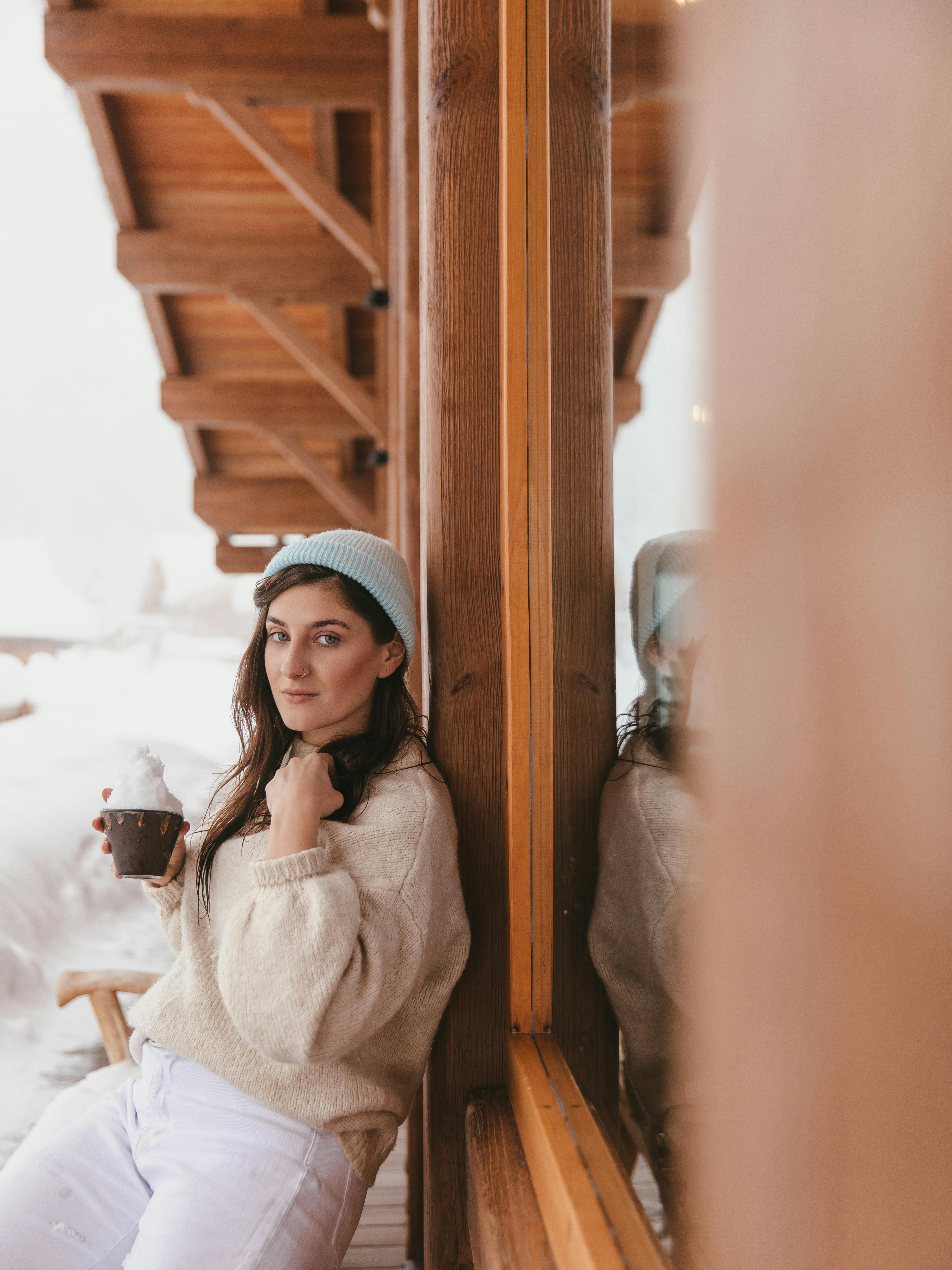 A woman in a beanie and sweater enjoys a hot drink while leaning on a cabin window in a snowy winter scene.