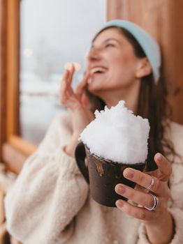 A joyful woman in a beanie holds a mug with snow, savoring a cozy winter moment indoors.