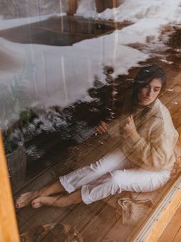 A woman sits reflectively indoors by a window, with a serene wintery scene outside.