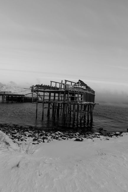 Black and white photo of a ruined pier on a snowy, rocky shore with a gloomy atmosphere.