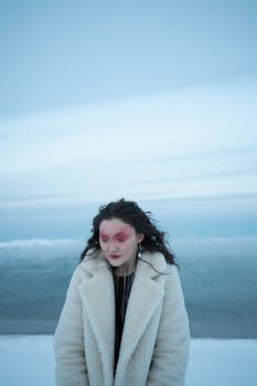Woman in a fur coat with artistic red makeup stands on a snowy beach during winter.