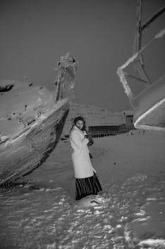 A woman stands amid snow-laden boats in a serene winter landscape.