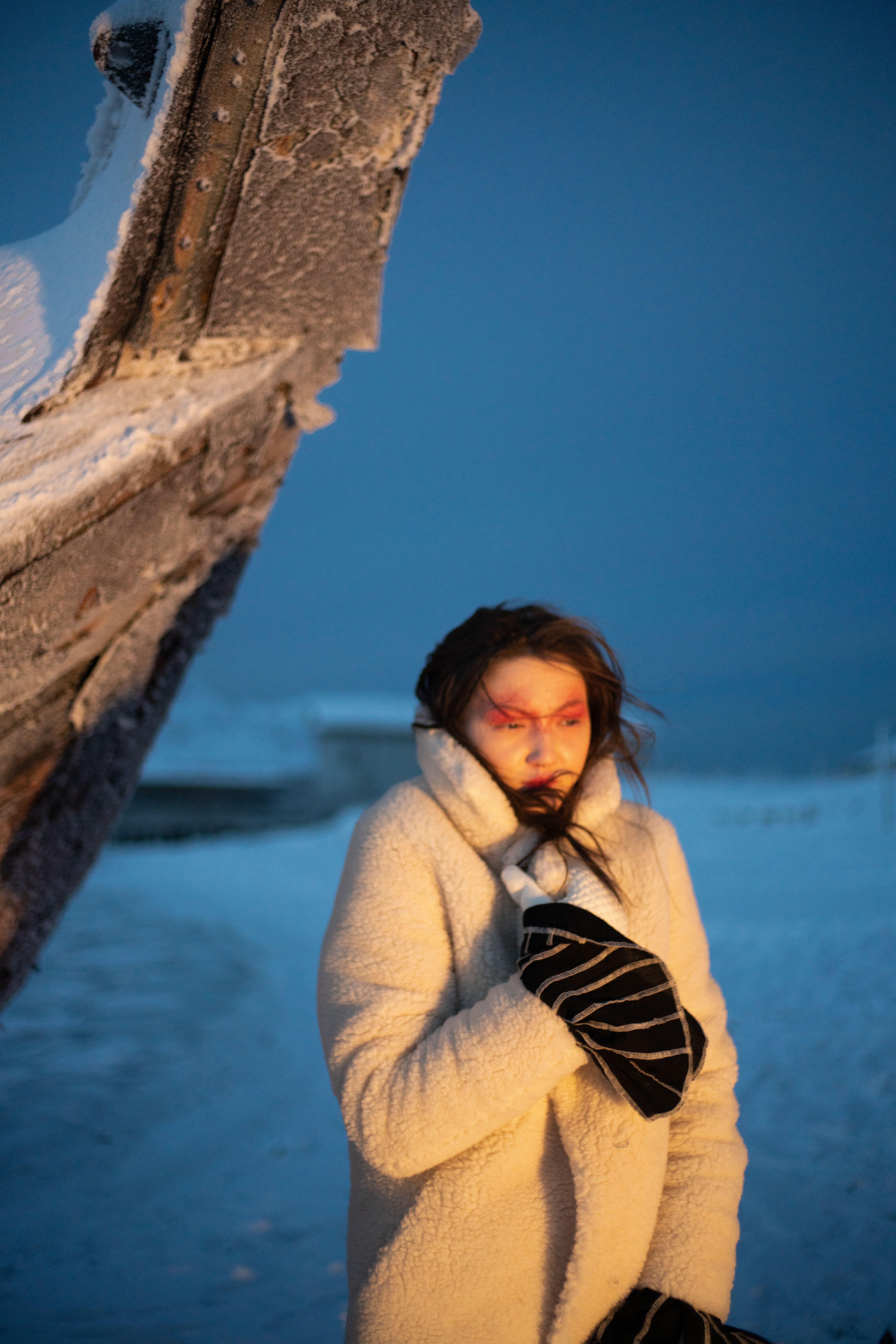 A woman in a winter coat stands amidst a snowy and icy landscape during the day, conveying a chilly atmosphere.
