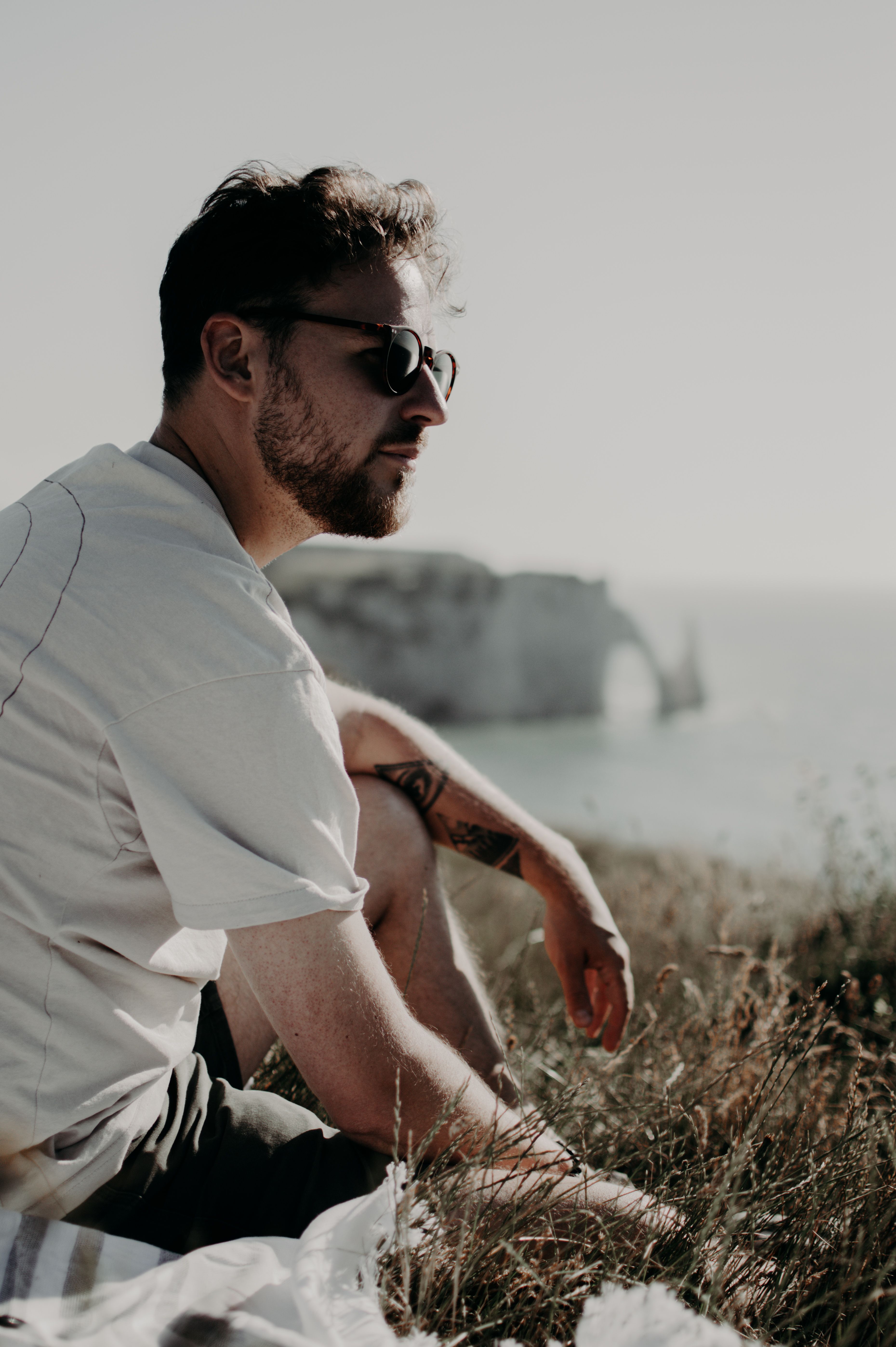 Side view of tattooed male in sunglasses sitting on plaid on grassy coast against rocky cliff near calm sea on blurred background