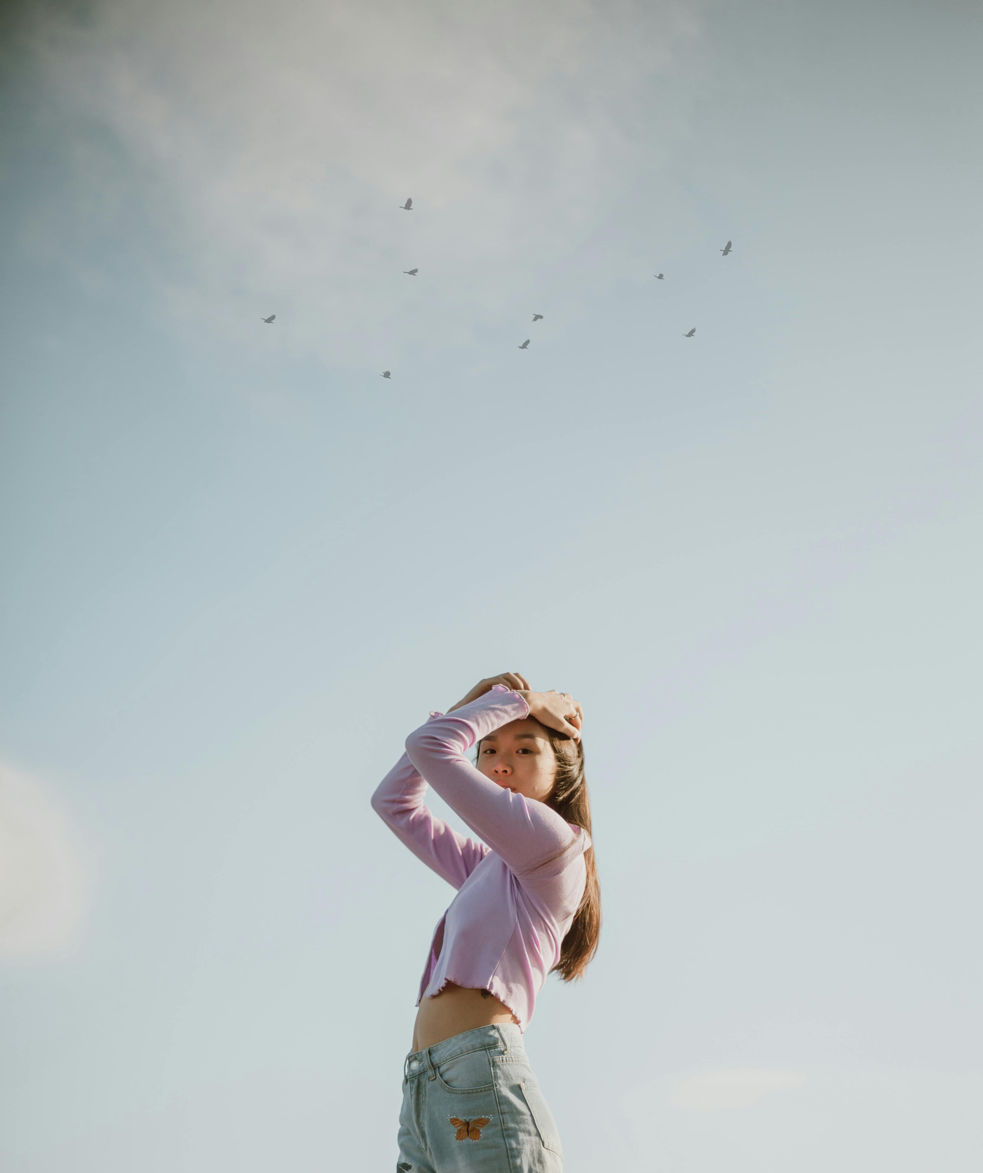 Young woman with hands on head looks up at the sky, surrounded by birds.
