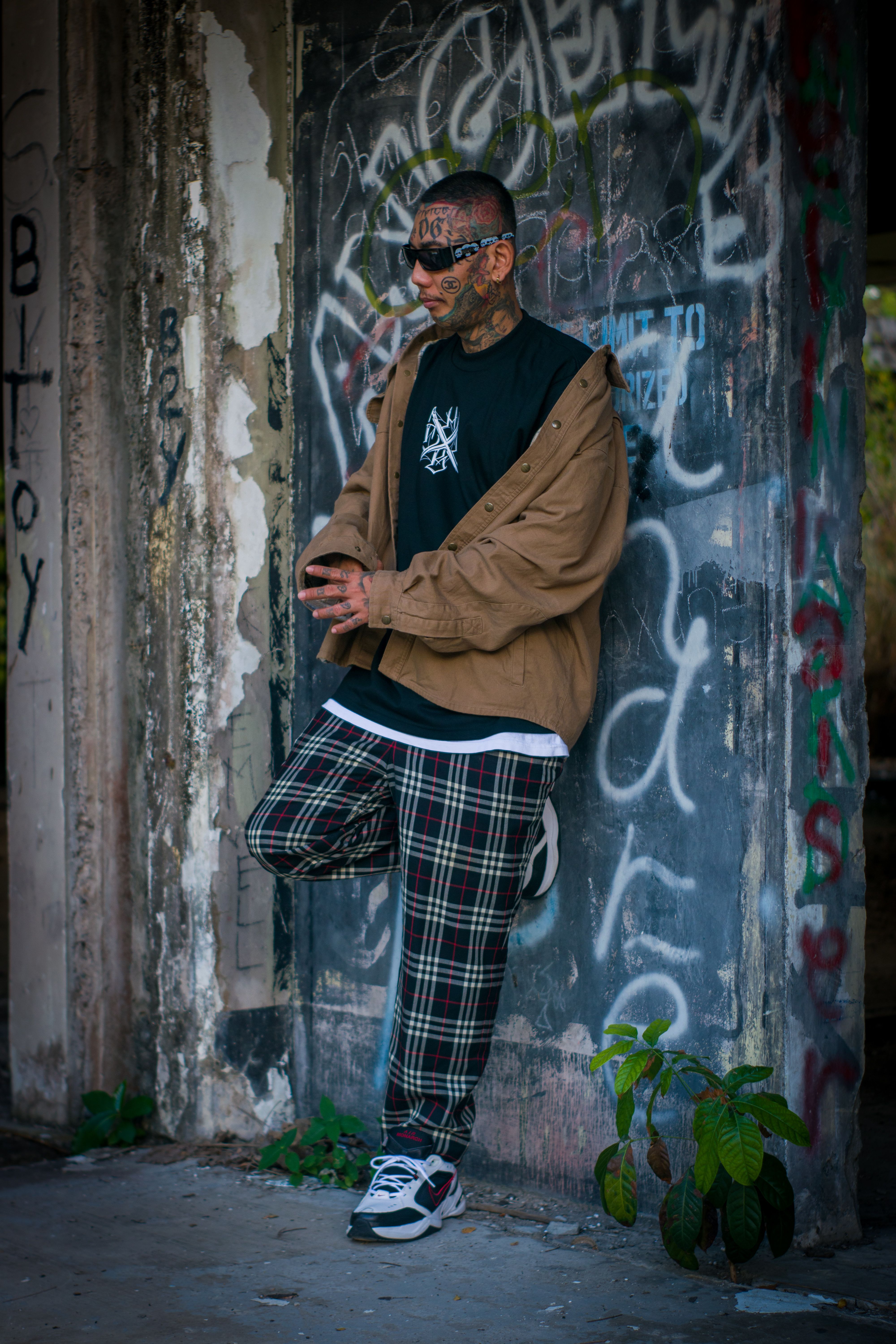 A man in plaid pants and tattoos stands against a colorful graffiti wall, exuding a relaxed urban vibe.