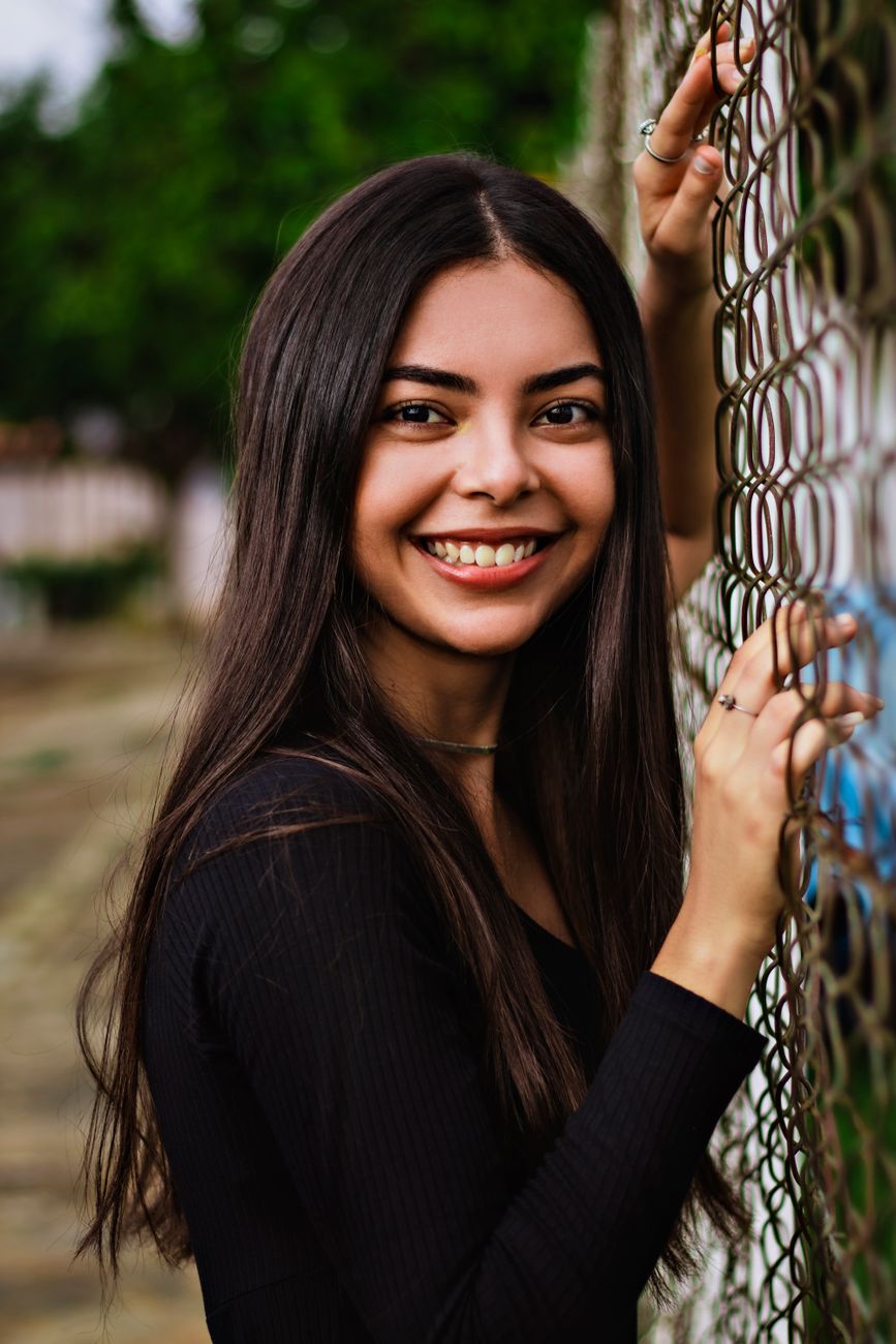 A young woman in black smiling and posing beside a chain-link fence outdoors.