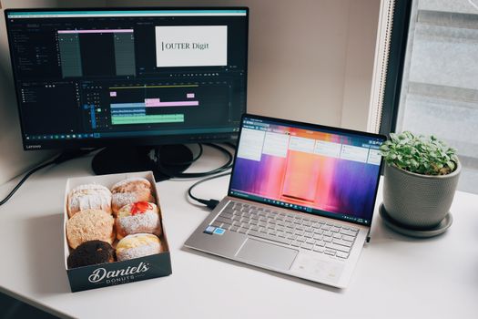 A workspace featuring a laptop, desktop monitor, box of donuts, and plant on a table.