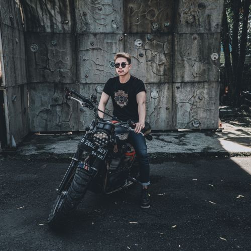 Free A fashionable man poses with his motorcycle against a textured wall on a sunny day, exuding confidence. Stock Photo