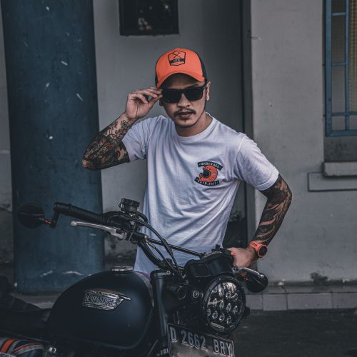 Free Tattooed man in sunglasses and cap posing by motorcycle outdoors, exuding style. Stock Photo