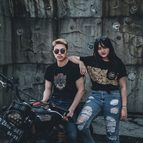 Free A trendy couple in casual attire posing confidently with a motorcycle against a weathered wall. Stock Photo