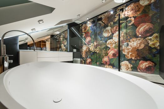 Oval shaped bathtub against shower room with floral ornament on wall in contemporary bathroom with lamps in house