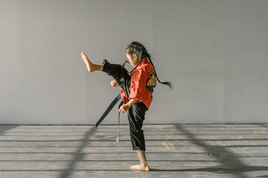 A young martial artist in a red uniform practicing a high kick indoors with natural lighting.