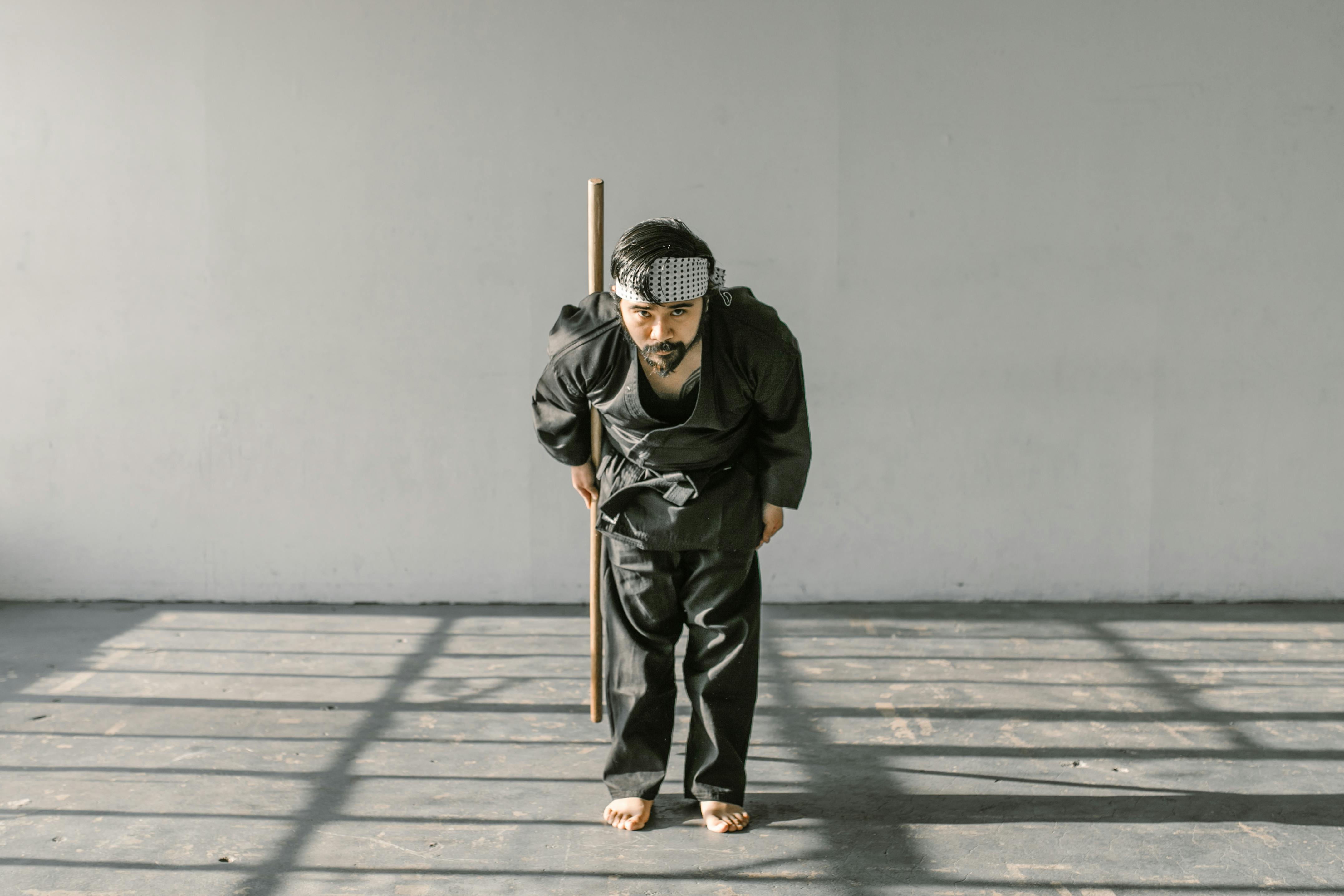 Free A martial artist in a black gi performs a traditional bow indoors with a wooden staff. Stock Photo