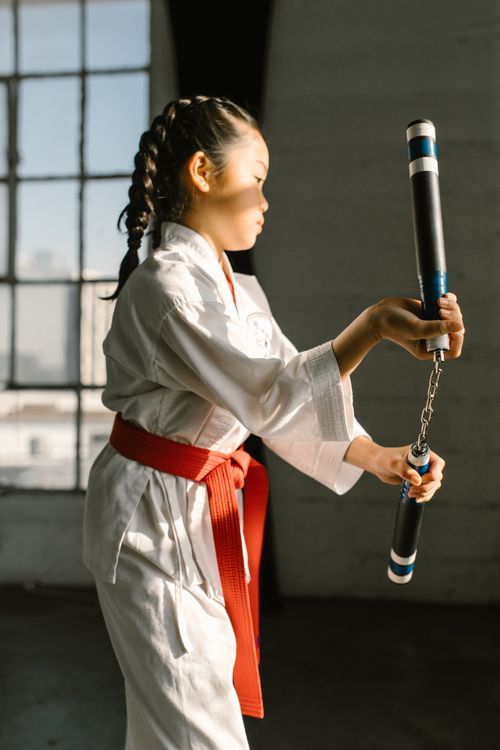 Free Asian girl practicing martial arts with nunchaku in a sunlit room. Stock Photo